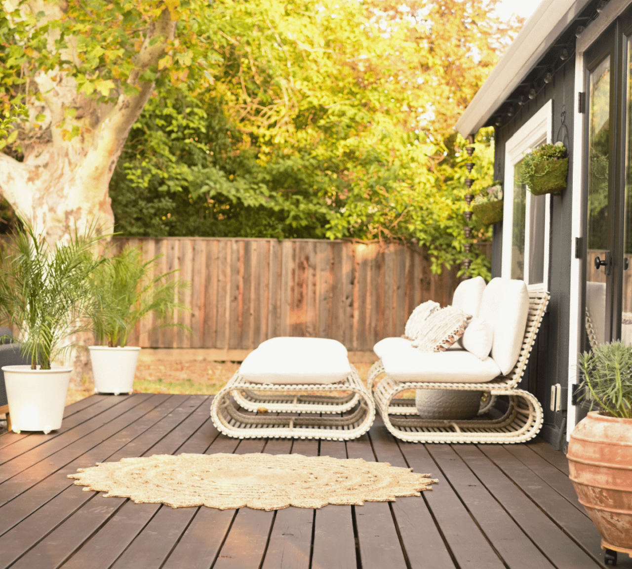 A deck stained a dark red-brown with potted plants, a circular outdoor rug and white modern furniture.