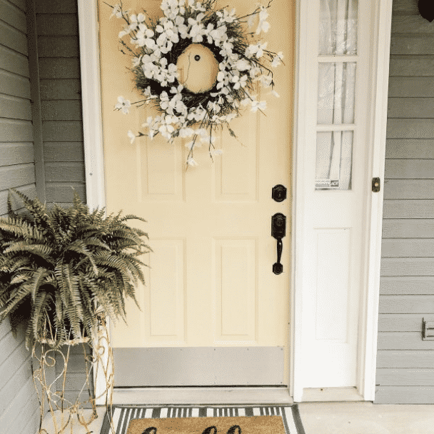 A front porch with welcome mat and a fern on a small table next to a light yellow front door. Photo credit to @sweetsouthernoaks.
